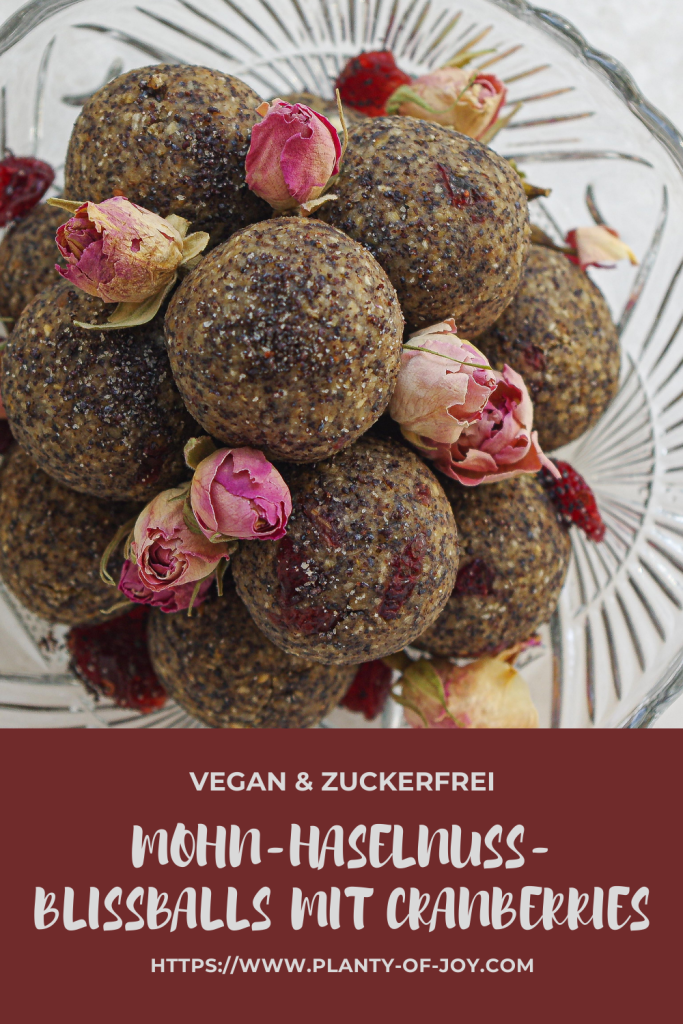 Mohn-Haselnuss-Blissballs mit Cranberries mit getrockneten Rosenblüten in einer Kristallglas-Schale auf einer weißen Steinplatte. Die Blissballs sind gestapelt und es wurden getrocknete Cranberries darüber gestreut. Der untere Teil des Bildes ist dunkelrot mit weißem Text. Der Text lautet in drei Zeilen vegan & zuckerfrei, Mohn-Haselnuss-Blissballs mit Cranberries und die url der Website Planty of Joy.