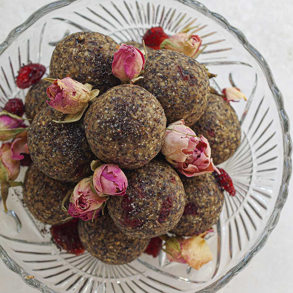 Mohn-Haselnuss-Blissballs mit Cranberries mit getrockneten Rosenblüten in einer Kristallglas-Schale auf einer weißen Steinplatte. Die Blissballs sind gestapelt und es wurden getrocknete Cranberries darüber gestreut.