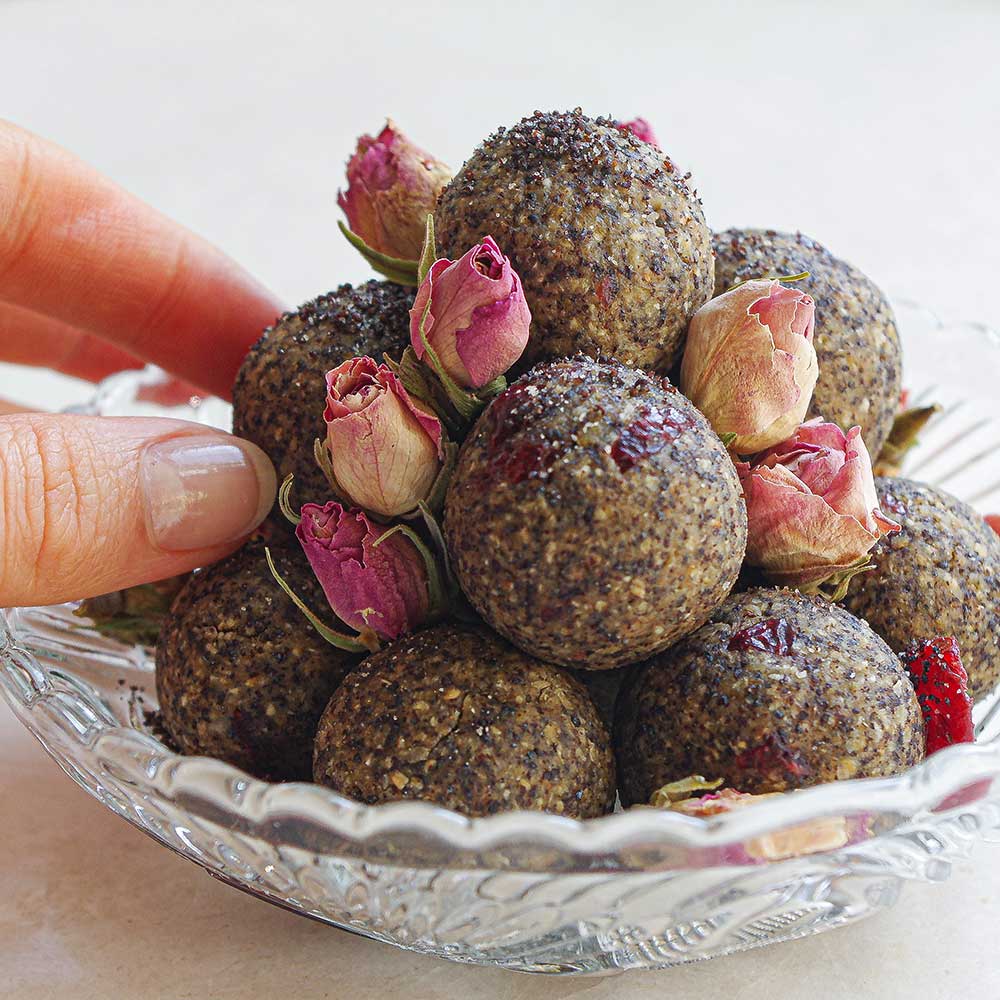 Mohn-Haselnuss-Blissballs mit Cranberries mit getrockneten Rosenblüten in einer Kristallglas-Schale auf einer weißen Steinplatte. Die Blissballs sind gestapelt und es wurden getrocknete Cranberries darüber gestreut. Eine Hand greift nach einem Blissball.