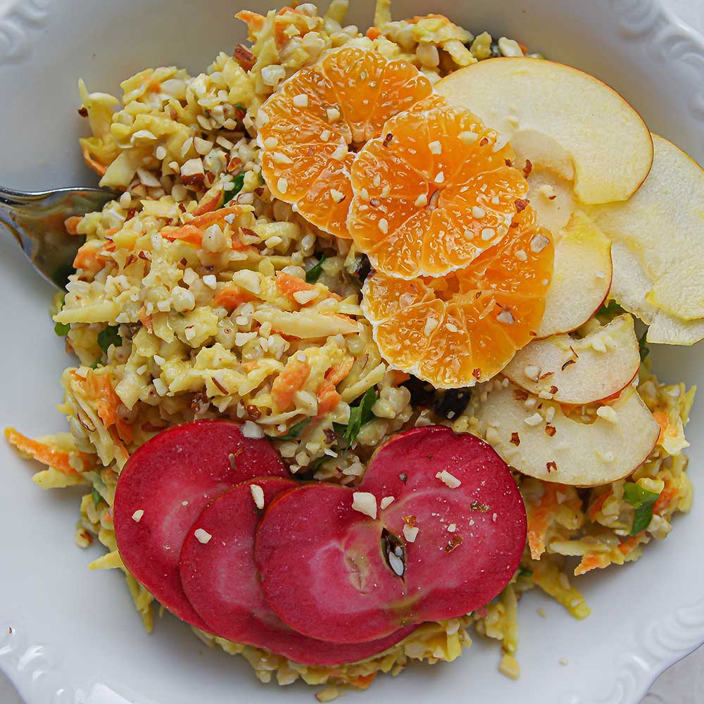 Eine Nahaufnahme von einer weißen Keramik-Schale mit gelbem Sellerie Salat mit Buchweizen und geraspelter Karotte. Der Salat ist getoppt mit Mandarinen-Scheiben, roten und hellgelben Apfelspalten. Die Schale steht auf einer weißen Steinplatte und neben der Schale liegt etwas frische Petersilie.