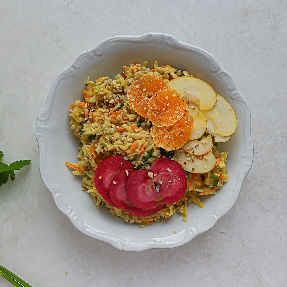 Eine weiße Keramik-Schale mit gelbem Sellerie Salat mit Buchweizen und geraspelter Karotte. Der Salat ist getoppt mit Mandarinen-Scheiben, roten und hellgelben Apfelspalten. Die Schale steht auf einer weißen Steinplatte und neben der Schale liegt etwas frische Petersilie.