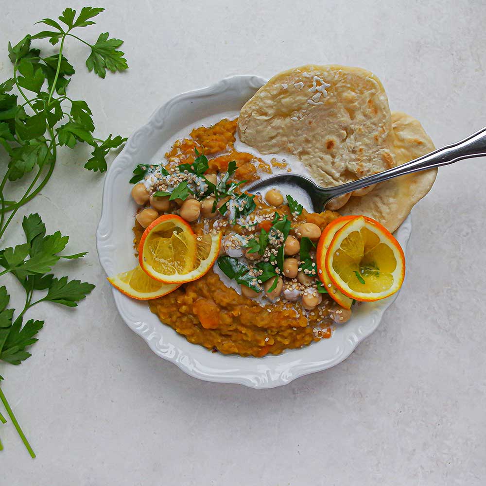 Eine helle Keramik-Schale gefüllt mit gelbem Linsen-Dal getoppt mit gehackter Petersilie, Kichererbsen und Naan.