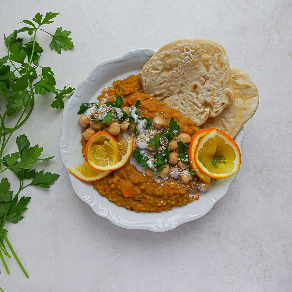 Eine helle Keramik-Schale gefüllt mit gelbem Linsen-Dal getoppt mit gehacktem Koriander. Im Hintergrund sieht man fluffiges Naan mit Feuerblasen und ein helles Geschirrtuch aus Leinen. Die Schale steht auf einem hellen Holztisch.