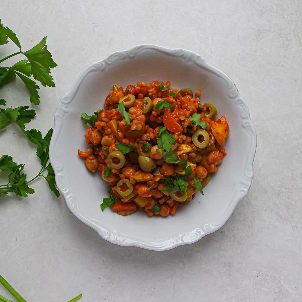 Mediterrane Couscous-Gemüsepfanne mit Oliven und frisch gehackter Petersilie in einer weißen Porzellan-Schale auf einer weißen Steinplatte mit frischer Petersilie daneben.