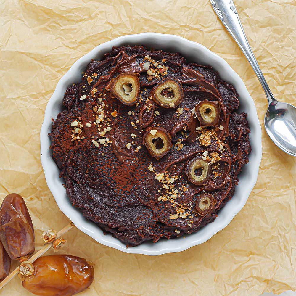 Vegane Schoko-Nuss-Creme in einer kleinen weißen Keramik-Schale getoppt mit klein geschnittenen Datteln, geschmolzener Schokolade und etwas Dattelzucker. Die Schale steht auf Backpapier.