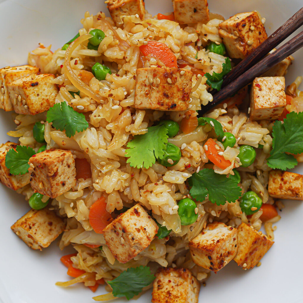 Eine Nahaufnahme von ebratenem Reis mit Gemüse getoppt mit Tofu und Koriander mit Holzstäbchen im Bild.