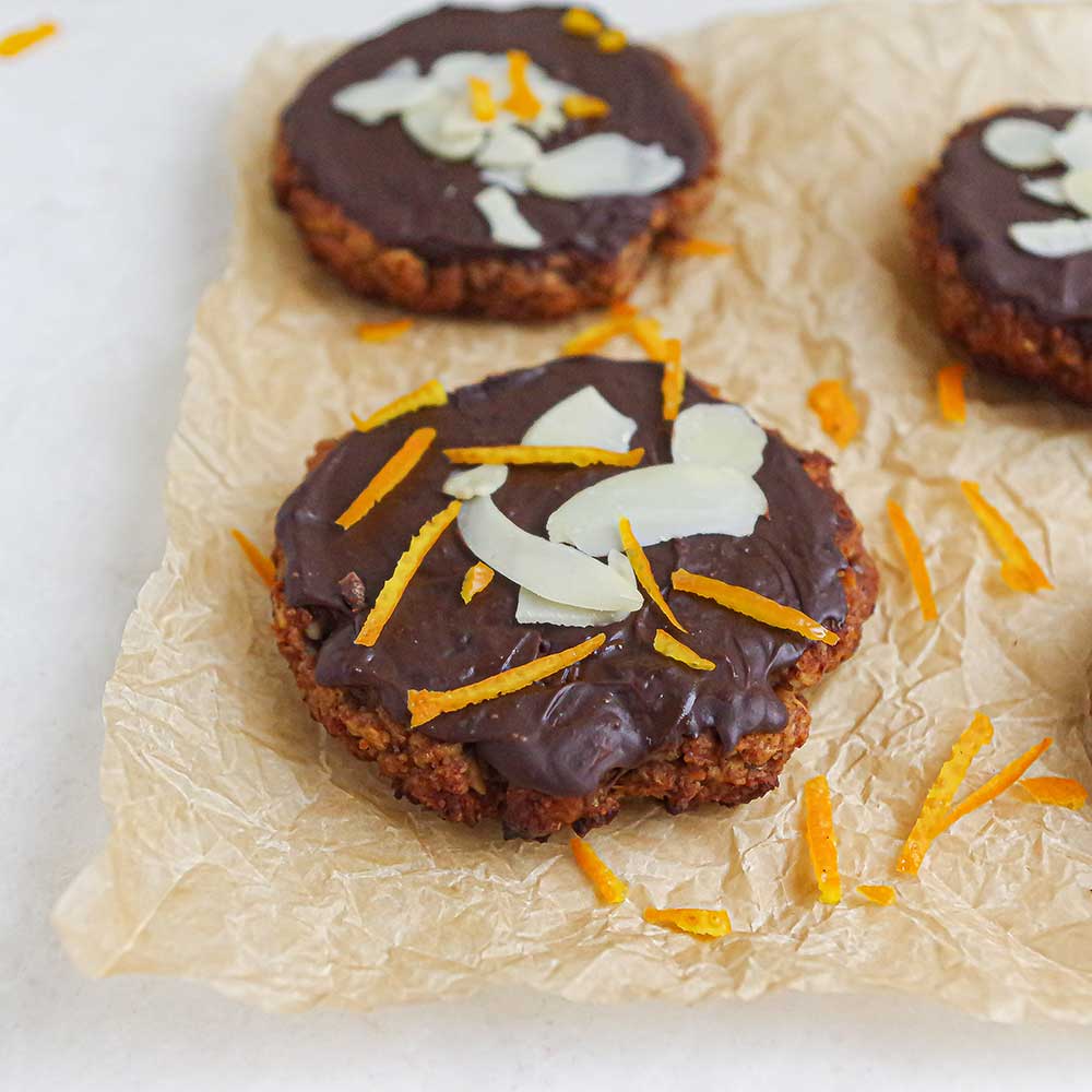 Leckere Mandel-Orangen-Cookies auf Backpapier. Die Cookies sind vegan, zuckerfrei und sind getoppt mit geschmolzener Schokolade, gehobelten Mandeln und dünnen Orangenschalen-Streifen.