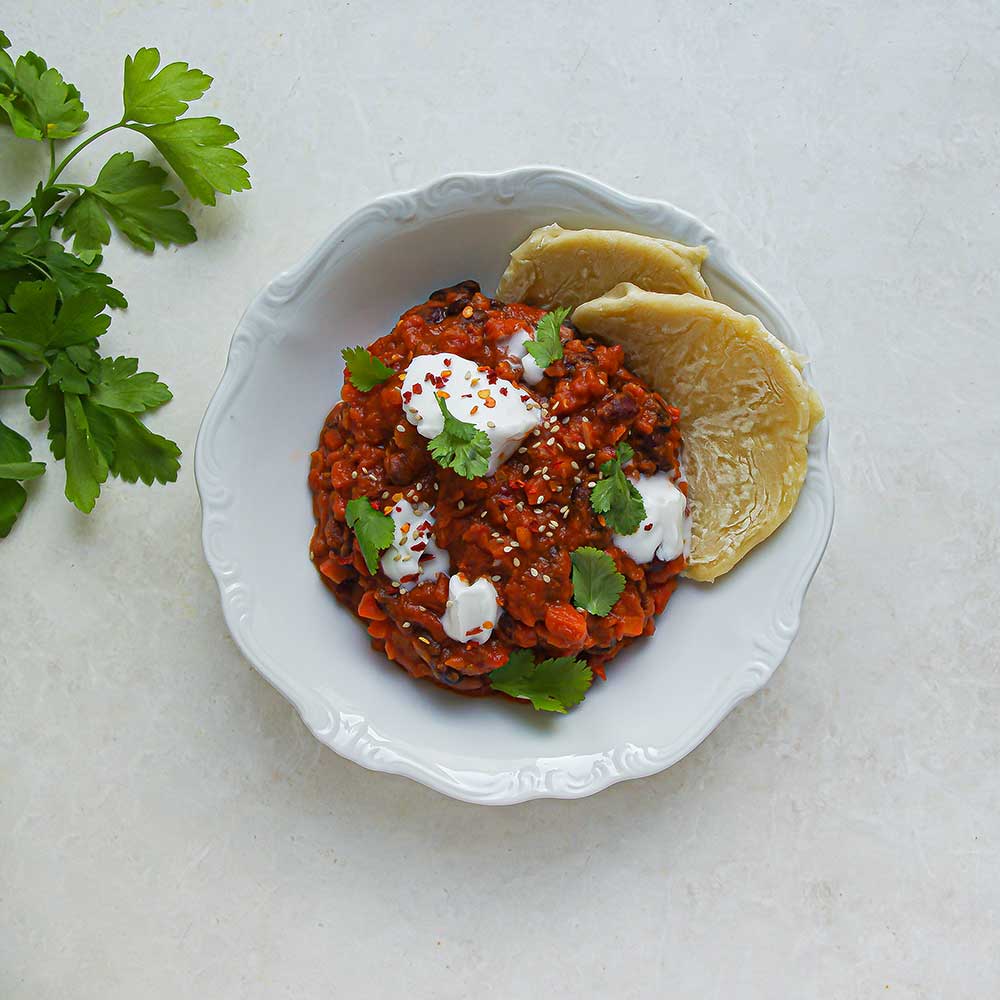 Proteinreiches Bohnen-Curry getoppt mit etwas Kokosjoghurt, Koriander und serviert mit Naan. Das Teller mit dem Curry steht auf einer weißen Steinplatte und daneben liegt frische Petersilie.