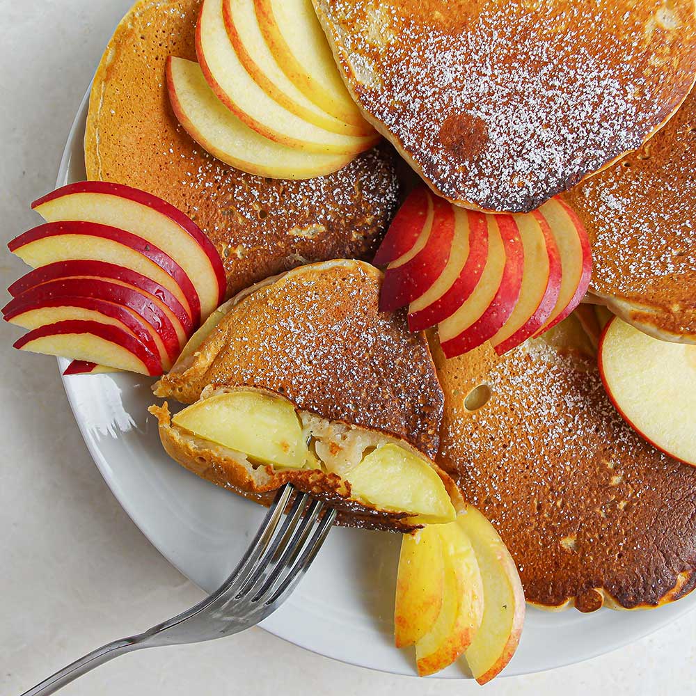 Gebackene-Apfel-Ringe mit Puderzucker bestäubt und dünn aufgeschnittenen Apfelspalten auf einem weißen Teller auf einer weißen Steinplatte.