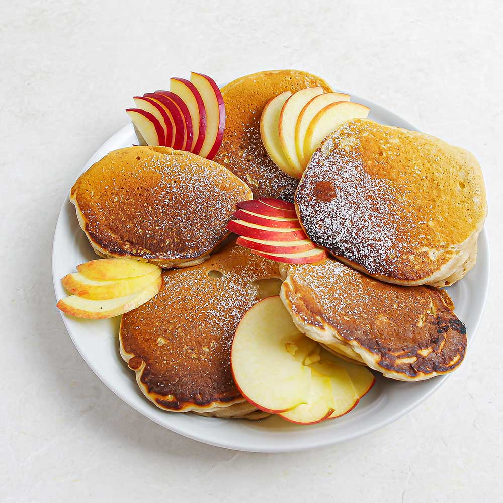 Gebackene-Apfel-Ringe mit Puderzucker bestäubt und dünn aufgeschnittenen Apfelspalten auf einem weißen Teller auf einer weißen Steinplatte.