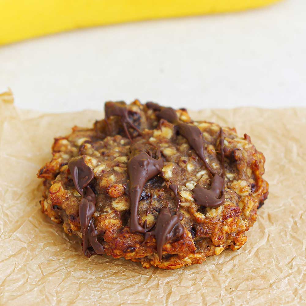 Ein brauner saftiger Bananenbrot-Cookie mit geschmolzener Schokolade verziert. Der Cookie liegt auf zerknittertem Backpapier. Im Hintergrund sieht man eine Banane.