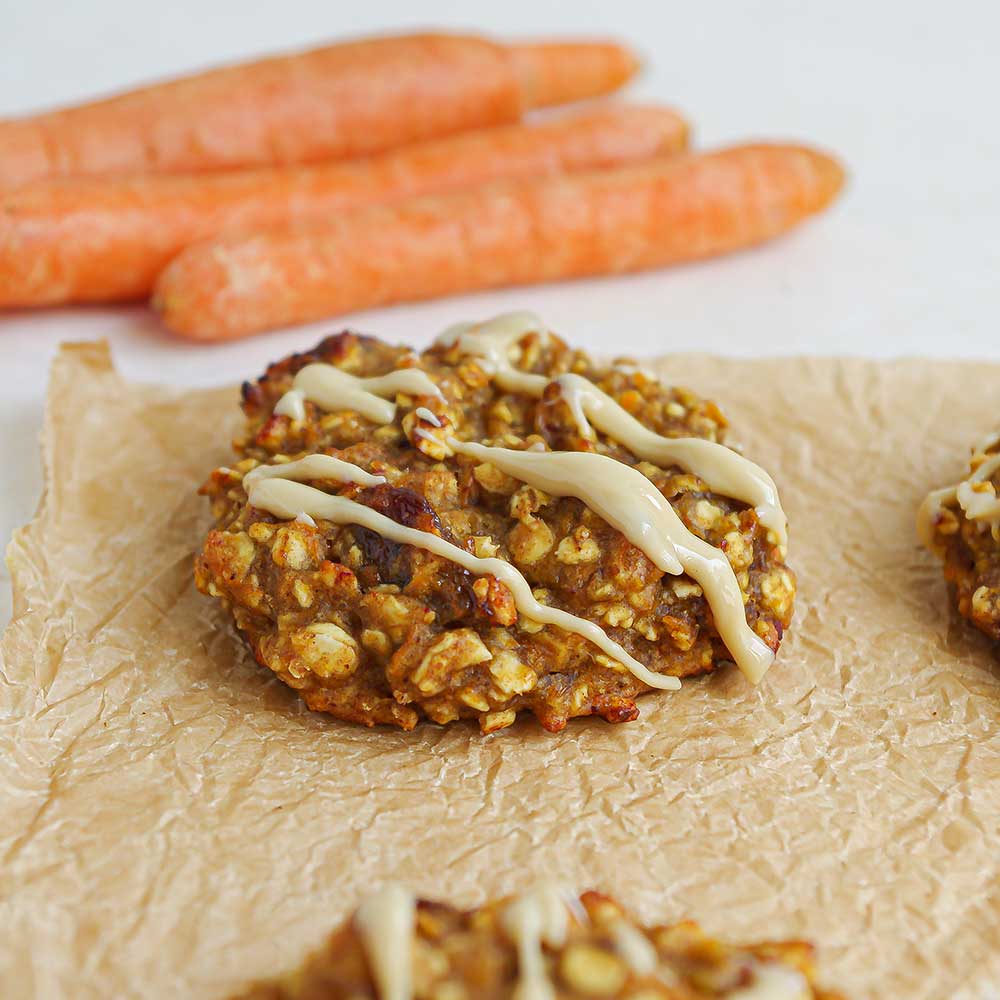 Eine Nahaufnahme eines saftigen Karotten-Hafer-Cookies mit Rosinen und Cashewmus-Drizzle. Der Cookie liegt auf zerknittertem Backpapier. Im Hintergrund sieht man Karotten und eine weiße Steinplatte.