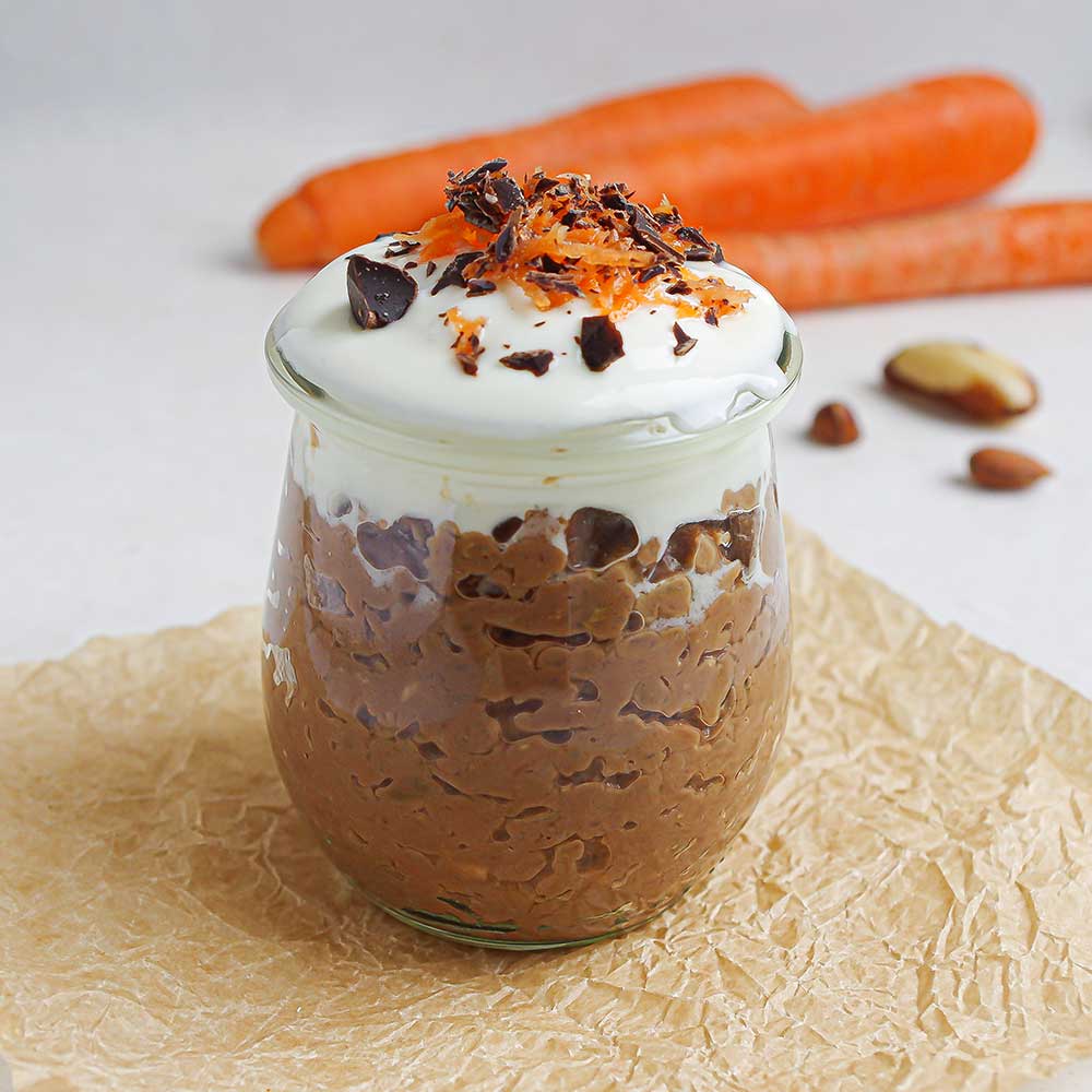 Ein kleines Weckglas gefüllt mit cremiger Schoko-Porridge mit Karotte getoppt mit Joghurt, geraspelter Karotte und gehackter Schokolade. Im Hintergrund sind Karotten und Nüsse. Das Weckglas steht auf zerknittertem Backpapier.