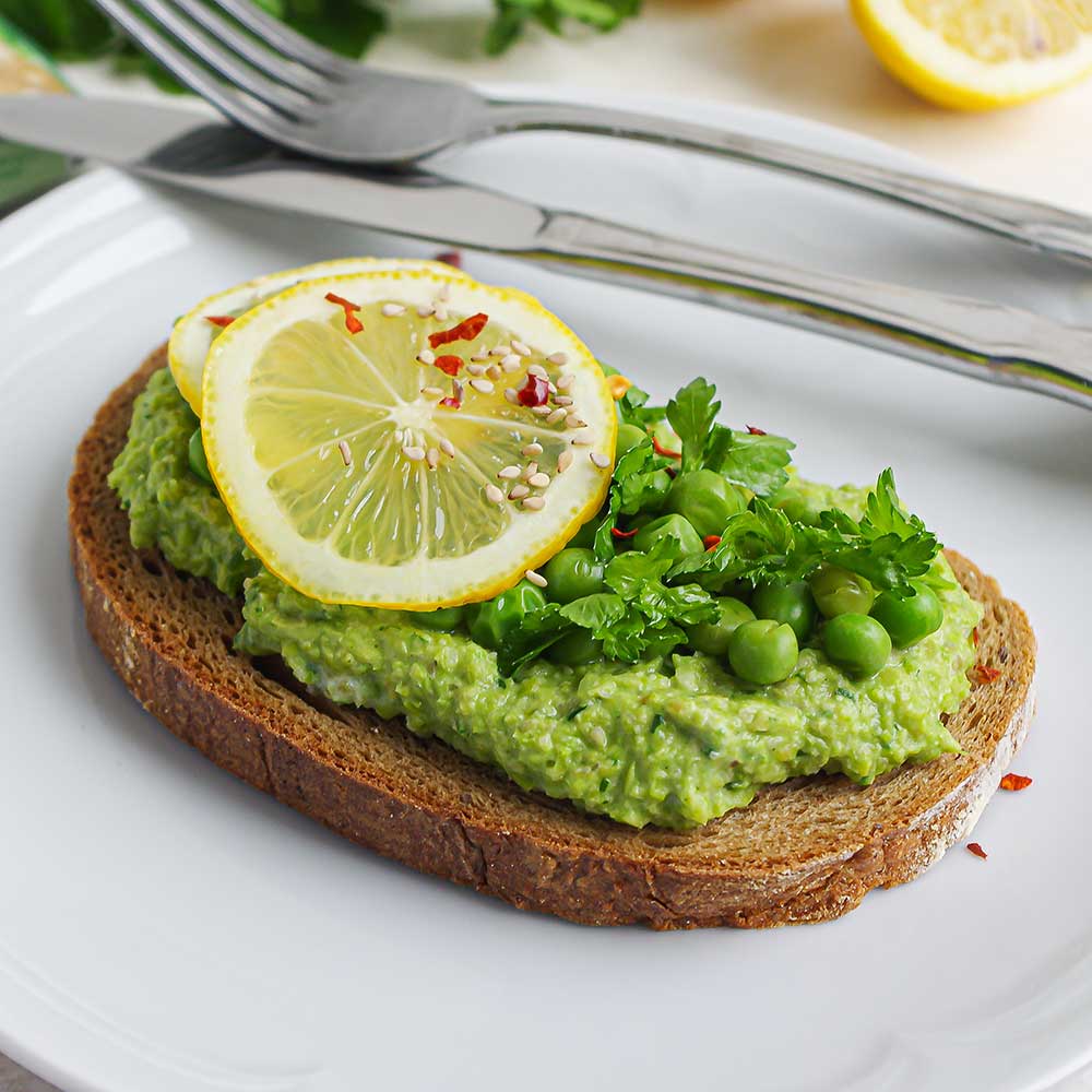 Ein Brot bestrichen mit grünem Erbsen-Kräuter-Dip getoppt mit Erbsen, gehackter Petersilie, zwei Zitronenscheiben und etwas Chili und Sesam. Das Brot liegt auf einem weißen Teller auf einer weißen Steinplatte und im Hintergrund sieht man frische Petersilie, eine halbe Zitrone und Messer und Gabel.