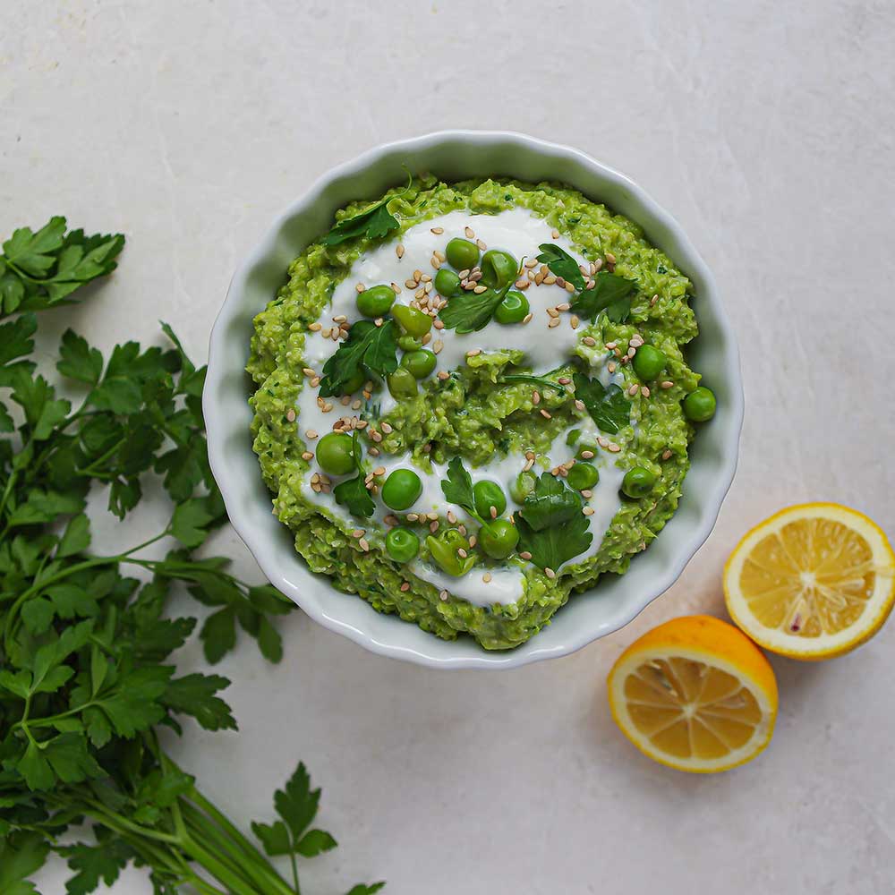 Eine weiße Keramik-Schale mit cremigem grünem Erbsen-Kräuter-Dip getoppt mit etwas Joghurt, Erbsen, gehackter Petersilie und Sesam. Die Schale steht auf einer weißen Steinplatte und im Hintergrund sieht man frische Petersilie und zwei halbe Zitronen.