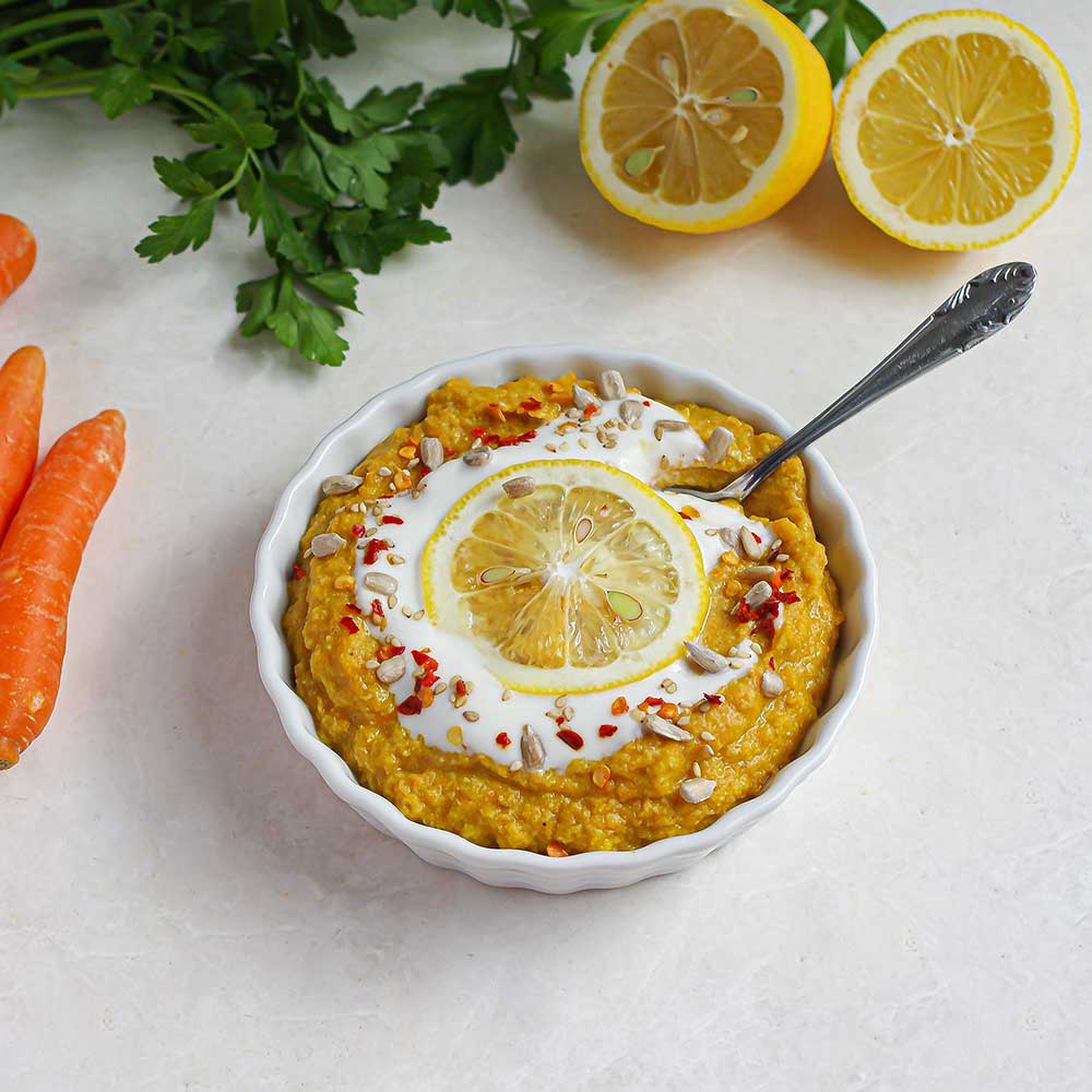 Eine weiße Keramik-Schale auf einer weißen Steinplatte mit dunkel gelbem Aufstrich, der mit einer Zitronenscheibe, Joghurt, Chiliflocken und Sonnenblumenkernen getoppt ist. Im Hintergrund sieht man halbe Zitronen, Karotten und frische Petersilie. Im Karotten-Linsen-Dattel-Dip steckt ein Löffel und man sieht wie cremig der Dip ist.