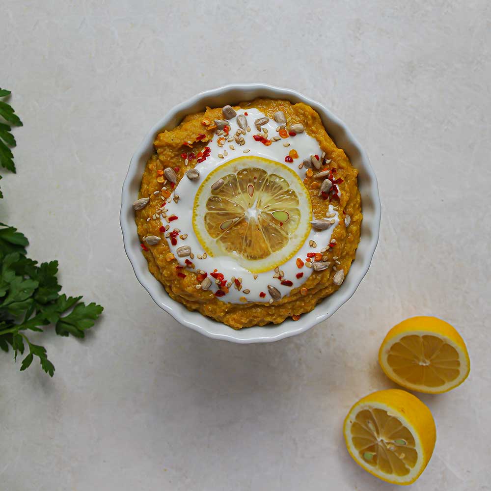 Eine weiße Keramik-Schale auf einer weißen Steinplatte mit dunkel gelbem Aufstrich, der mit einer Zitronenscheibe, Joghurt, Chiliflocken und Sonnenblumenkernen getoppt ist. Im Hintergrund sieht man eine halbe Zitrone und frische Petersilie.