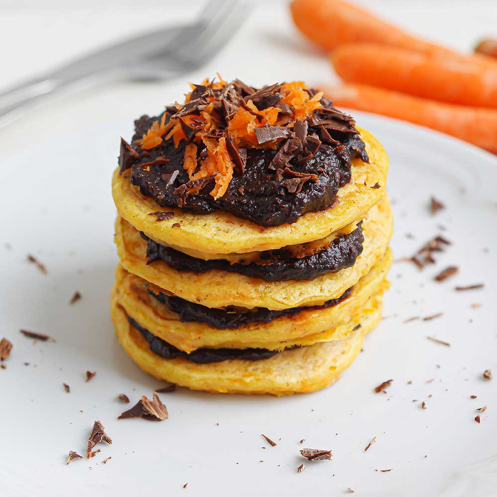 Fluffige Karotten-Haselnuss-Pancakes gestapelt mit Schoko-Creme dazwischen und getoppt mit Schoko-Creme und geraspelter Karotte. Im Hintergrund sind Karotten und Besteck zu sehen.