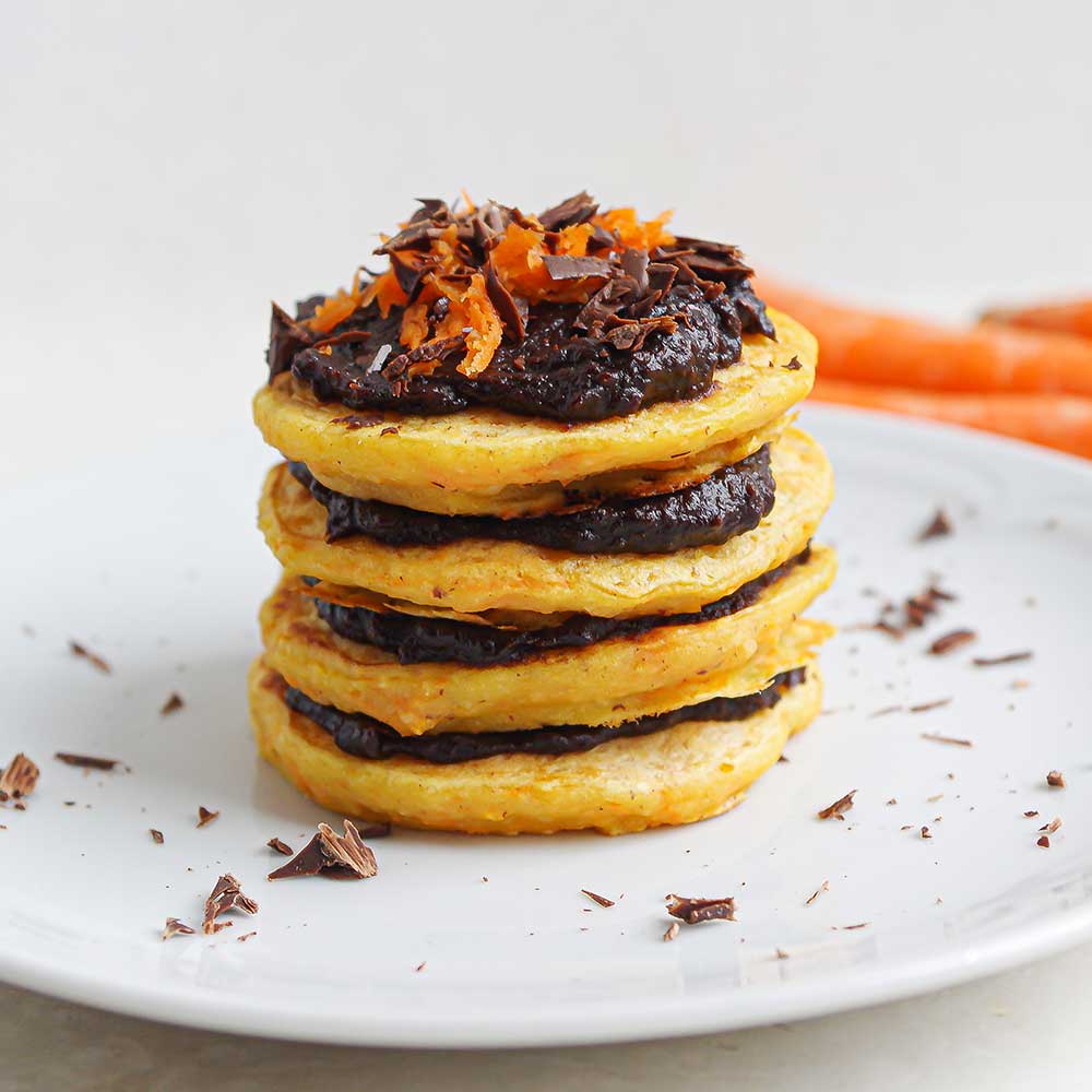 Fluffige Karotten-Haselnuss-Pancakes gestapelt mit Schoko-Creme dazwischen und getoppt mit Schoko-Creme und geraspelter Karotte. Im Hintergrund sind Karotten zu sehen.