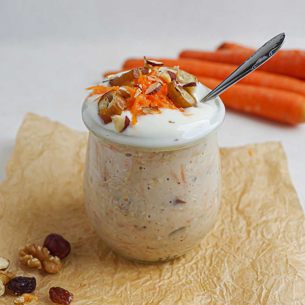 Karottenkuchen-Birchermüsli in einem kleinen Weckglas getoppt mit Joghurt, geraspelter Karotte, Datteln und gehackten Haselnüssen. Das Weckglas steht auf zerknittertem Backpapier auf einer weißen Steinplatte und im Hintergrund sind Karotten und ein paar Nüsse zu sehen.