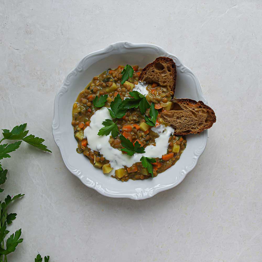 Eine weiße Keramikschale gefüllt mit veganem Berglinsen-EIntopf getoppt mit knusprigem Brot, Joghurt und frisch gehackter Petersilie. Die Keramikschale steht auf einer weißen Steinplatte mit frischer Petersilie im Hintergrund.