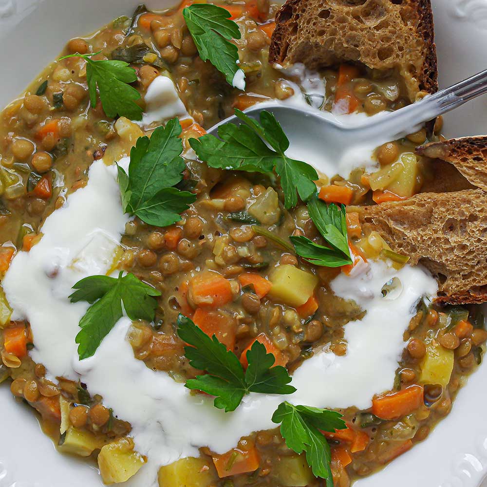 Eine Nahaufnahme von veganem Berglinsen-EIntopf getoppt mit knusprigem Brot, Joghurt und frisch gehackter Petersilie.