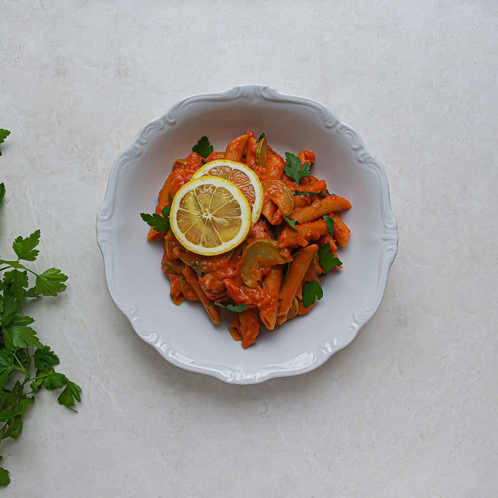 Eine weiße Keramik-Schale gefüllt mit cremiger Tomaten-Pasta getoppt mit zwei Zitronenscheiben und gehackter Petersilie. Die Schale steht auf einer weißen Steinplatte und am Rand sieht man etwas frische Petersilie.