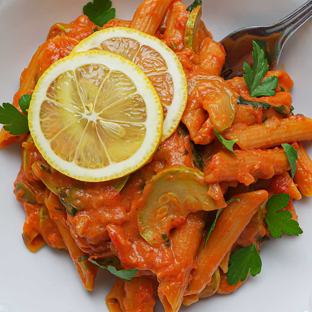 Eine Nahaufnahme einer weißen Keramik-Schale gefüllt mit cremiger Tomaten-Pasta getoppt mit zwei Zitronenscheiben und gehackter Petersilie.