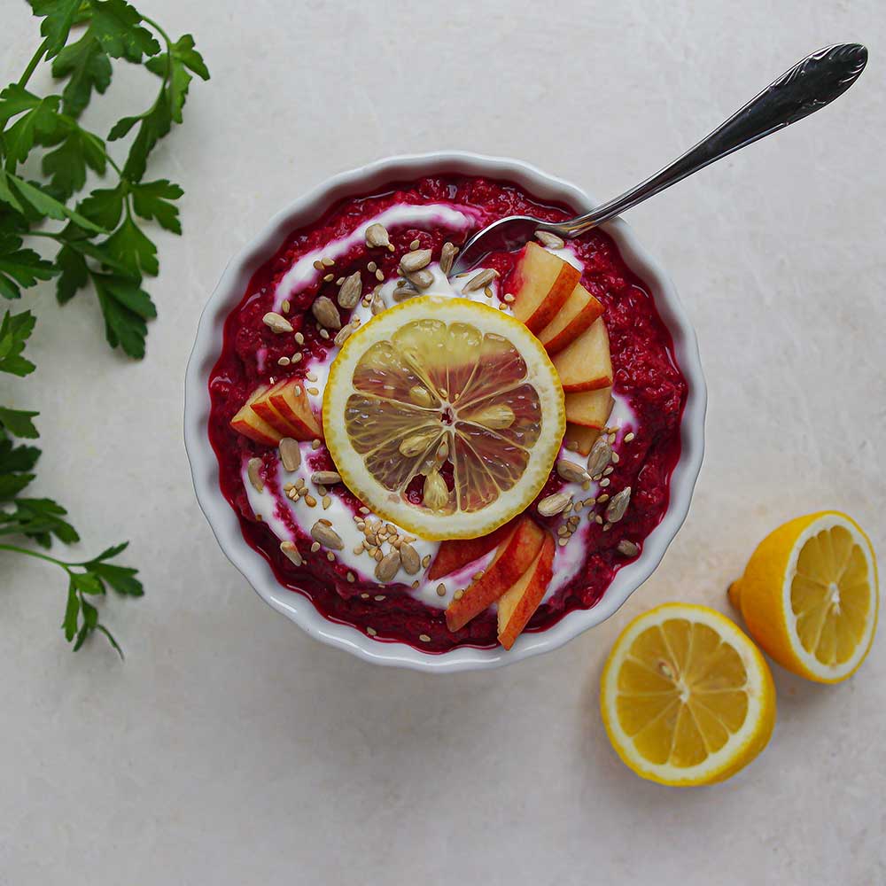 Eine weiße Keramik-Schale mit Pinke Rote Rüben-Apfel-Dip getoppt mit Joghurt, Apfelstückchen, einer Zitronenscheibe und Sonnenblumenkernen. Im Dip steckt ein Löffel und der Dip steht auf einer weißen Steinplatte mit zwei Zitronenhälften und etwas frischer Petersilie im Hintergrund.