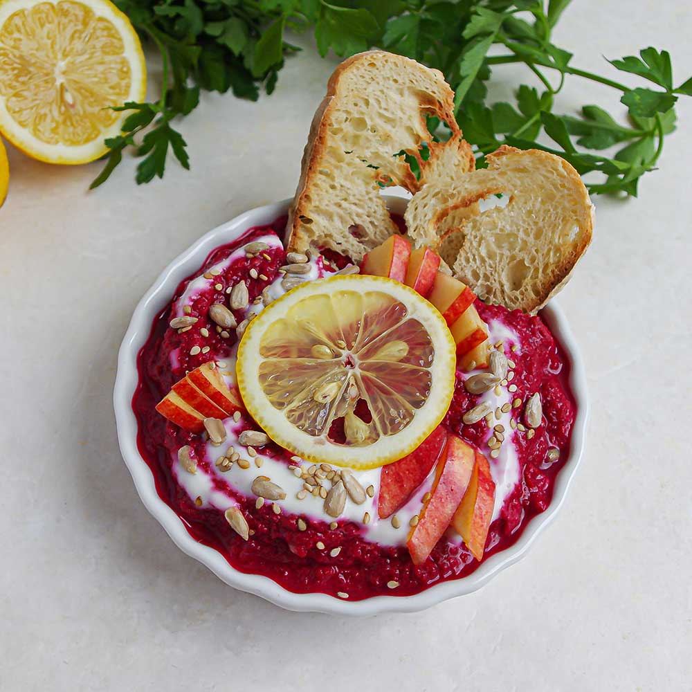 Eine weiße Keramik-Schale mit Pinke Rote Rüben-Apfel-Dip getoppt mit Joghurt, Apfelstückchen, einer Zitronenscheibe und Sonnenblumenkernen. Im Dip stecken zwei knusprige Brotstückchen und der Dip steht auf einer weißen Steinplatte mit zwei Zitronenhälften und etwas frischer Petersilie im Hintergrund.