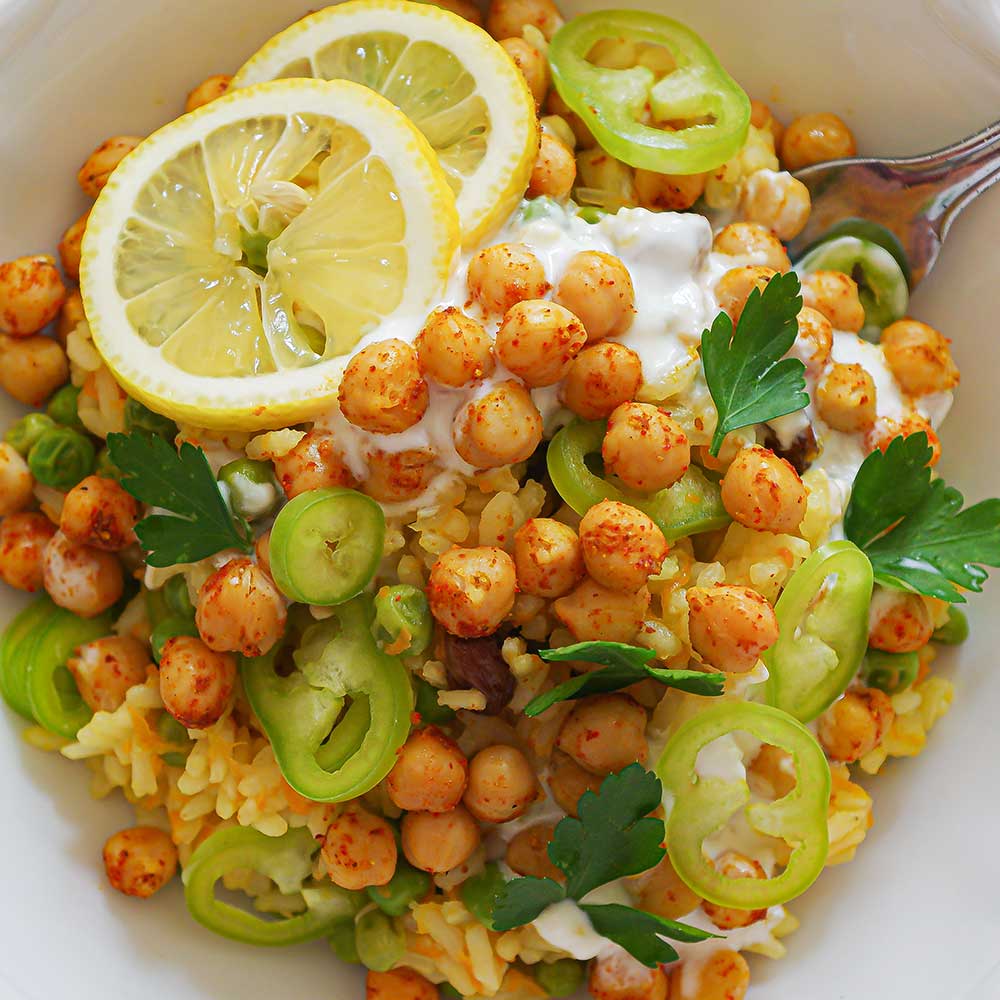 Eine Nahaufnahme von gelbem Safranreis getoppt mit Paprika, Zitronenscheiben, Joghurt, Petersilie und gerösteten Kichererbsen.