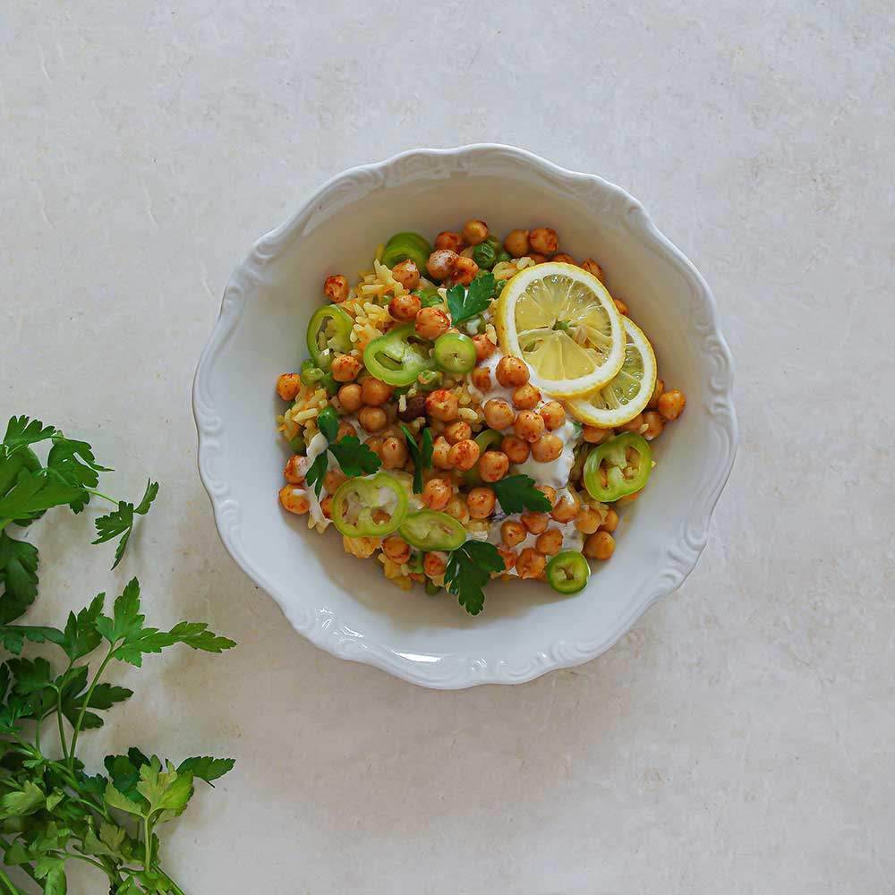 Gelber Safranreis getoppt mit Paprika, Zitronenscheiben, Joghurt, Petersilie und gerösteten Kichererbsen. Der Reis wird in einer weißen Keramik-Schale serviert, die auf einer weißen Steinplatte steht. Neben der Schale liegt frische Petersilie.