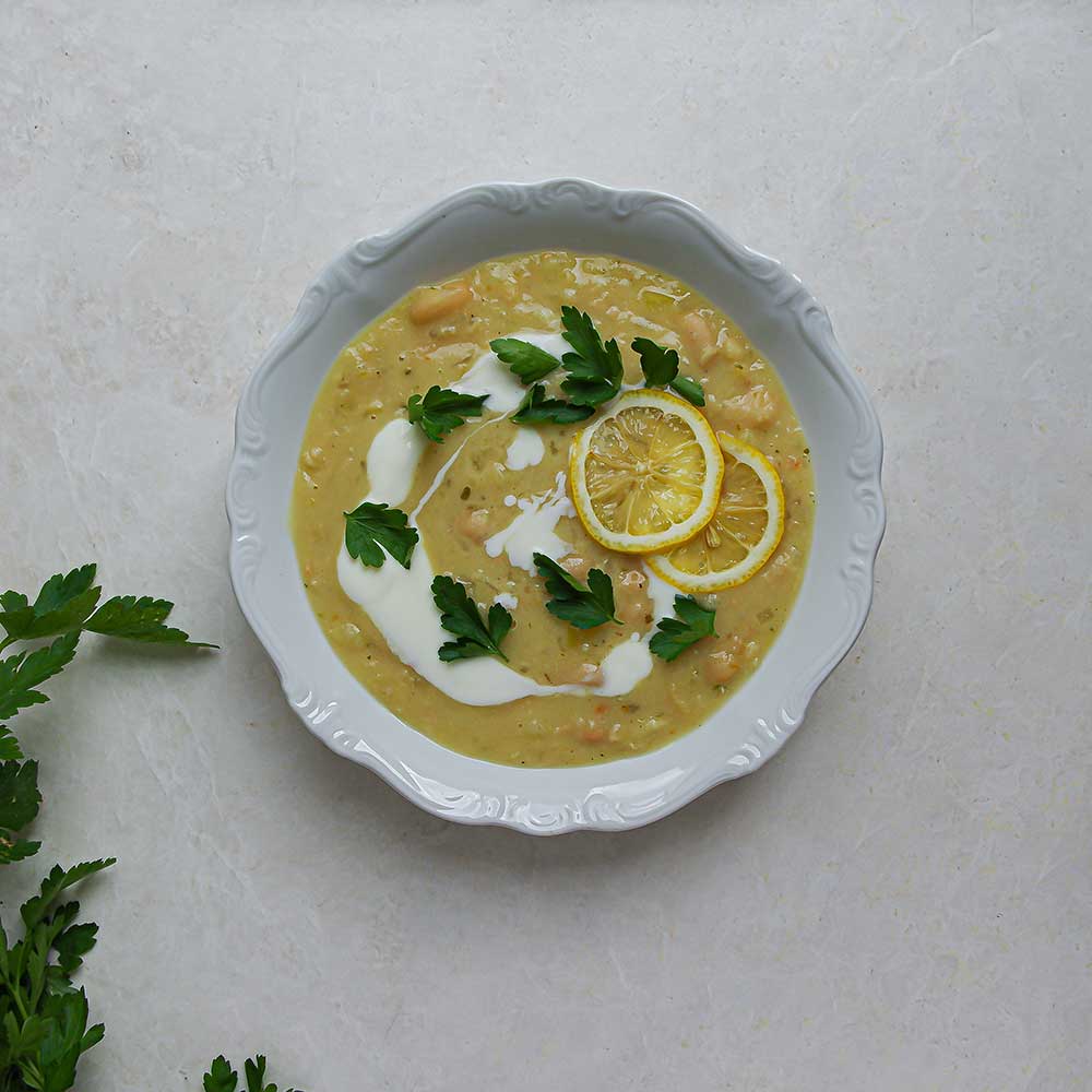 Cremige Weiße Bohnen-Zitronen-Suppe in einer weißen Keramik-Schale getoppt mit frischer Petersilie, Joghurt und einer Zitronenscheibe. Die Schale steht auf einer weißen Steinplatte mit frischer Petersilie im Hintergrund.