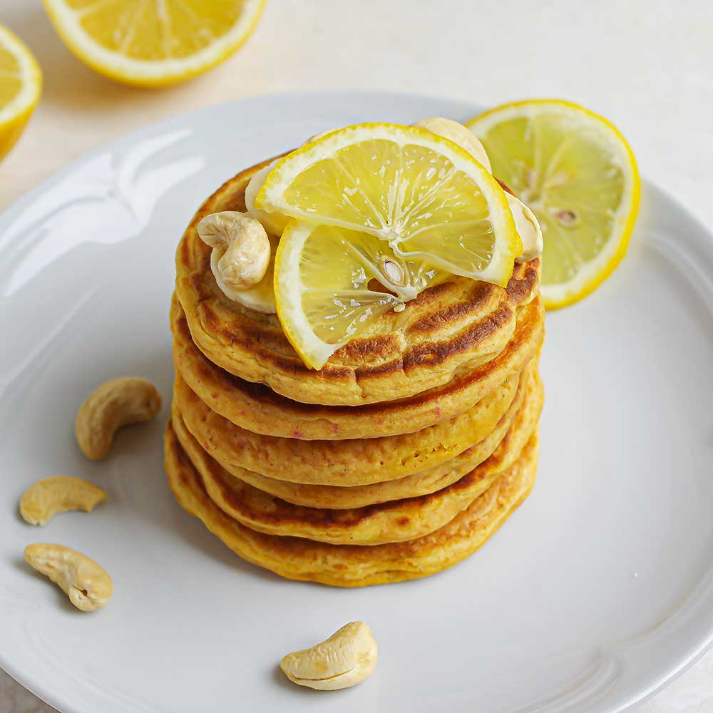 Fluffige Zitronen-Pancakes getoppt mit Zitronenscheiben und Cashews auf einem weißen Keramikteller. Im Hintergrund sind halbe Zitronen. Die Pancakes sehen sehr saftig und lecker aus und haben eine gold-gelbe Farbe.