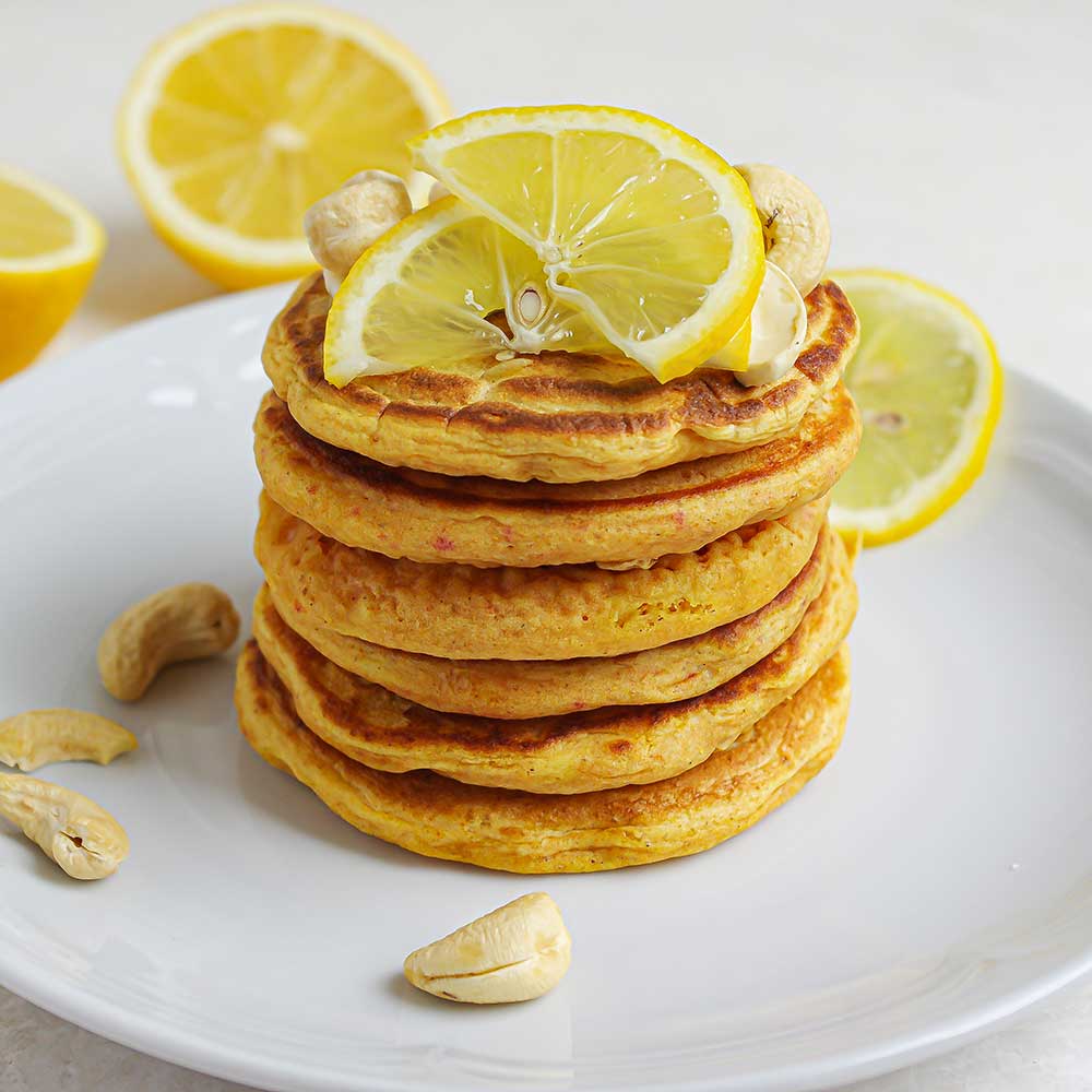 Fluffige Zitronen-Pancakes getoppt mit Zitronenscheiben und Cashews auf einem weißen Keramikteller. Im Hintergrund sind halbe Zitronen. Die Pancakes sehen sehr saftig und lecker aus und haben eine gold-gelbe Farbe.