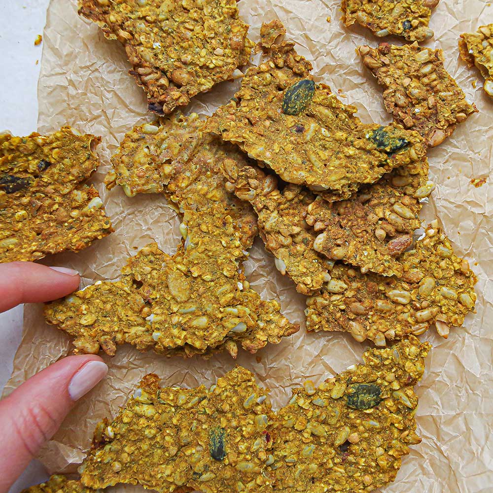 Curry-Kurkuma-Cracker auf Backpapier. Die Curry-Kurkuma-Cracker sehen sehr knusprig und lecker aus mit vielen Saaten, Kernen und Samen.