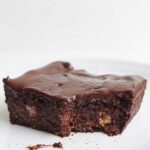 Ein Bild von veganen fudgy Brownies mit Schokoladenglasur und getoppt mit Himbeeren. Der Brownie ist quadratisch und serviert auf einem kleinen Keramik-Teller.