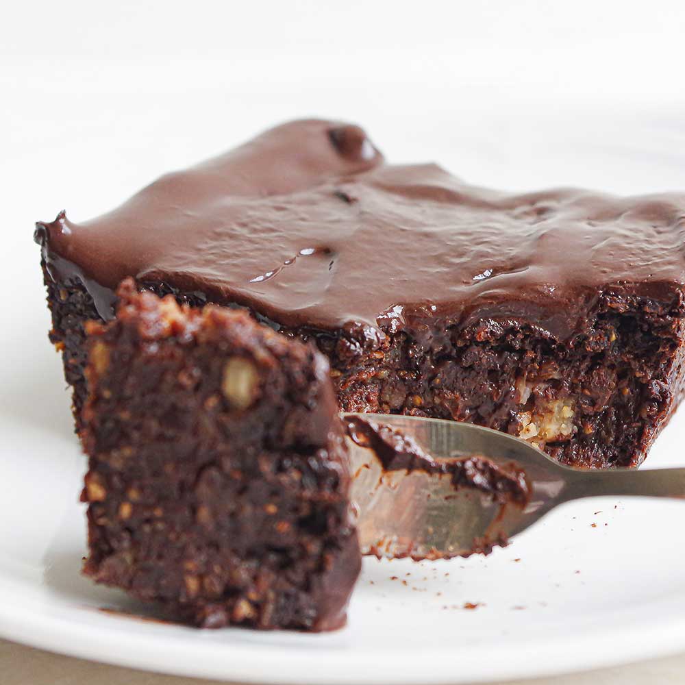 Ein Bild von veganen fudgy Brownies mit Schokoladenglasur und getoppt mit Himbeeren. Der Brownie ist quadratisch und serviert auf einem kleinen Keramik-Teller.