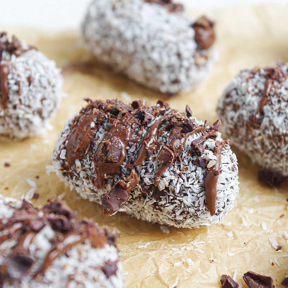 Protein-Schoko-Kokos-Riegel auf zerknittertem Backpapier als Nahaufnahme. Die Riegel sind in Kokosraspeln gewälzt, mit Schokolade verziert und mit Kakaonibs bestreut.