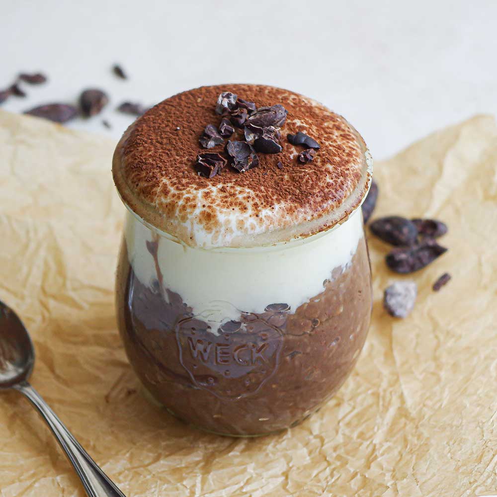 Schokoladiger Tiramisu-Porridge geschichtet mit veganem Joghurt und bestäubt mit Back-Kakao und Kakaonibs in einem kleinen Weckglas.