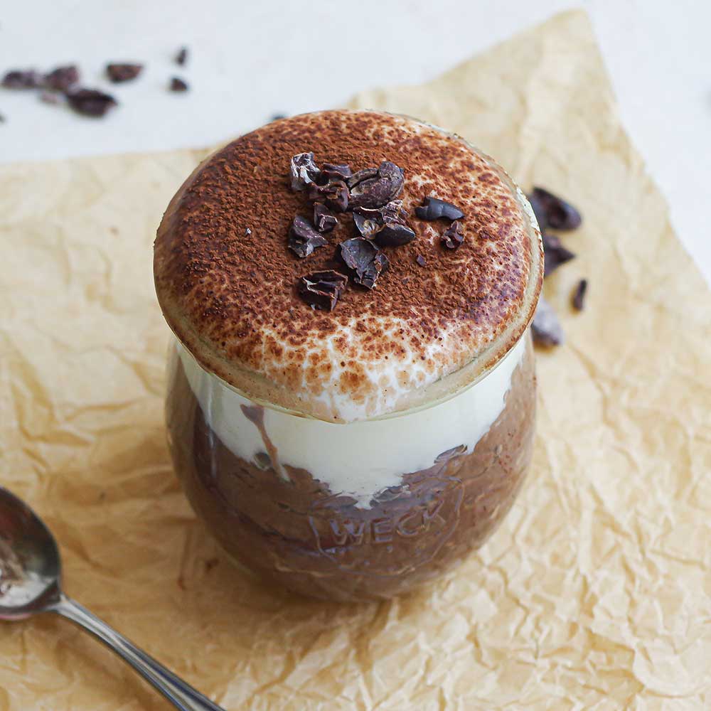 Schokoladiger Tiramisu-Porridge geschichtet mit veganem Joghurt und bestäubt mit Back-Kakao und Kakaonibs in einem kleinen Weckglas.