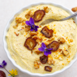 Nahaufnahme eines cremigen Dattel-Curry-Topfen-Dips in einer weißen Schüssel, mit Walnüssen, Datteln, Blüten und einem Löffel.